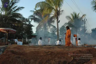 A photograph of an outdoor homa fire site on reddish earth, with smoke drifting across the scene. A long-haired figure in saffron-orange robes walks at right, holding a folded orange cloth in one hand. Several men in white ritual clothes sit or crouch around the smouldering fire pit, and another in white stands behind them. A white and red striped ceremonial umbrella stands at left, with coconut palms and a green-roofed building behind. The clothing and setting suggest a fire ritual in progress.