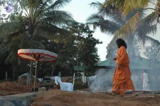 A photograph of an outdoor ritual site on dusty red earth beside coconut palms and green-roofed sheds. In the centre-right, a figure in long orange robes and sandals walks past, with long dark hair swept back, a hand raised in gesture. Smoke rises from a fire area behind him. On the left, a white and red fringed canopy stands on a pole above a white-draped table. A second person in white clothing stands in the right background.