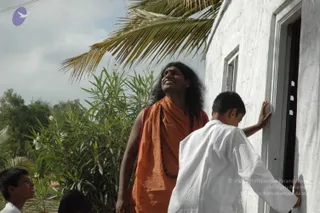 Outdoor photo beside a whitewashed wall with a window and doorway, under a palm frond and green oleander bushes. A long-haired man with a red tilak on his forehead wears an orange-saffron shawl over his shoulders and a rudraksha mala, and looks upward with an open-mouthed expression. Beside him a boy in white clothes places his hand on the wall near the doorway. Two more children's heads are partly visible at the lower left edge. The man's long hair, saffron wrap and beads are visible cues for Swamiji (probable), shown as a person standing outdoors.
