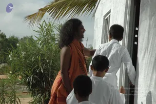 A photograph outdoors beside a white rendered building with a doorway and window. A man with long, swept-back dark hair wears an orange-saffron wrap and a long rudraksha mala, and smiles as he reaches toward the door. Three boys in white kurta-style clothes stand near the entrance, one reaching for the door frame. A leafy oleander shrub with white flowers and a palm frond are in the background under a cloudy sky. The visible physical people are the man and three boys.