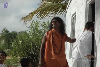 A photograph outdoors beside a whitewashed building with a palm frond overhead and green oleander bushes. A man with long, swept-back dark hair and a red mark on his forehead wears an orange saffron wrap over his shoulders, a rudraksha mala around his neck, and a red cloth beneath. He smiles and rests one hand on the doorframe of the white building. To the right, a person in white clothes leans out of the doorway. At the lower left, two young boys are partly visible at the frame's edge.
