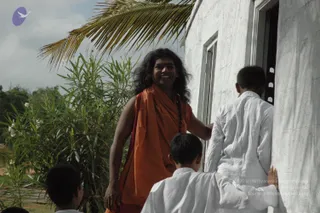 Outdoor photograph beside a whitewashed building with a dark doorway. A man with long, swept-back dark hair and a red bindi stands at center in a saffron-orange shawl over an orange garment, with a rudraksha mala visible at his neck, smiling and holding the doorframe. Three boys in white kurtas stand around him: one at the right entering the doorway, one bottom right with hand on the wall, and a partly visible child at bottom left. A palm frond and green shrubs with white flowers frame the scene under a pale sky.