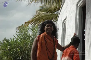 A smiling man with long, dark, swept-back hair wears a saffron-orange shawl over his shoulders and a dark rudraksha mala around his neck, standing outdoors beside a whitewashed wall with a window. His right hand rests on the head of a young boy in an orange shirt who is partly visible at the right edge. Behind them are palm fronds and green oleander bushes under an overcast sky.