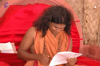 A photograph of a long-haired man in an orange saffron shawl and rudraksha mala seated against a bright red fabric backdrop with a woven straw wall to the right. He has a red tilak on his forehead, looks down and smiles while reading a printed sheet held in both hands. a person is shown in an appearance-based label: long swept hair, orange saffron shawl and rudraksha mala. A second person's hand and printed paper appear at the far left edge.