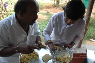 Two men in plain white shirts and white trousers sit under an open shelter eating rice from stainless steel plates. The man on the left, with a moustache and a red-thread wristband, points at his plate holding yellow rice and a flat pappadum-like cracker. The younger man on the right, with long dark hair, leans over his own plate of rice with a spoon. Behind them are dry earth, green grass, a wooden post and a green plastic container. No a person candidate is visible.