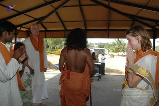 A photo taken under an open thatched-roofed pavilion with palm trees beyond. A figure with long dark hair seen from behind, bare-backed, wearing orange ochre cloth wrapped around the body and holding a walking cane, stands facing a group. On the left, a young man in white with an orange shawl presses palms together in anjali. Next, an older man with glasses in a white kurta and orange shawl stands with hands at his sides. On the right, a woman with light hair in a white sari with gold border and an orange shawl stands with palms pressed together in namaskar. A small table and people in white appear in the background, and a parked vehicle is visible beyond the pavilion.