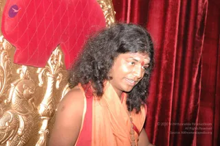 A photograph of one man seated on an ornate gold throne with a red velvet cushion, in front of a red velvet curtain. He has long, dark, wavy hair swept back, a red tilak mark on his forehead, and wears an orange-saffron shawl over a red garment with a long beaded necklace of gold-toned pendants. Swamiji (probable): long swept-back hair, saffron clothing and a ceremonial necklace on a throne, consistent with the page's Deepavali event at Aadhi Peetha, Bidadi. The image shows a seated person with a brass-tone throne carving of a lion-like figure to the left.
