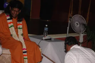 A photograph of a seated figure in a saffron-orange robe with a multicoloured flower garland of white, pink, orange, green and navy blossoms draped over the chest, smiling and looking down. A man in a white shirt and white trousers bends forward at the lower right to touch the seated figure's feet. A white-draped table holds a water bottle and a microphone, a white pedestal fan stands beside it, and a red and dark stage backdrop with gold-edged curtains fills the background.