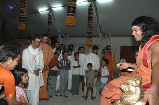 A photograph inside a tent-like hall with white fabric canopy and hanging decorative cloth panels in orange, yellow and black patterns. On the right, a long-haired man in orange and red robes with a red-and-white tilak on his forehead sits and gestures with an open hand toward a group of children. A golden lion-shaped object sits in front of him. Standing in a semicircle are about a dozen boys in white kurtas and dhotis, a boy in a red shirt, and a boy in a checked shirt, plus a man in a white dhoti and shawl with a red lanyard badge and a bearded man in orange robes. At left, a woman in an orange sari and a young girl in pink are visible. A cameraman in orange robes stands behind. a person appearance is an uncertain candidate, so this is marked unsure.