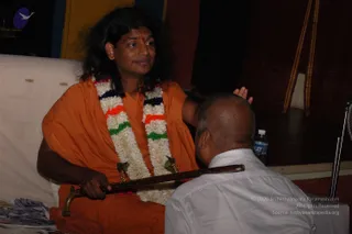 A seated man in an orange-saffron robe wears a white and green-orange-navy flower garland around his neck and a red tilak on his forehead. His long dark hair falls past his shoulders. He rests one hand on the head of a bald man in a white shirt who bows forward toward him. The seated man holds a long wooden staff with a metal-ended handle across his lap. Behind them are a white cushioned seat, a water bottle, and a dark draped backdrop with yellow-striped fabric. Stacks of printed booklets sit at the lower left. The scene matches the Deepavali blessing described on the page. a person is seated, identifiable by his long swept hair, saffron robe and floral garland, and is giving blessings.