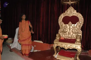 Indoor photo in a hall with deep red velvet drapes. A man with long dark swept-back hair, wearing an orange saffron robe with red border, a long rudraksha mala, bare feet and an upraised hand, stands on a red-carpeted platform beside an ornate gilded wooden throne with carved birds and owl-like lion figures, a red velvet seat and backrest, and a hanging gold canopy. Behind him a man in white clothes stands, and at left a man in orange holds a red tray with sweets.