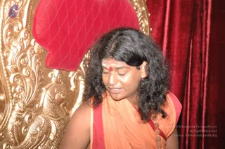 A photograph of a long-haired man in an orange and red saffron wrap, with a red kumkum mark and white vibhuti on his forehead, seated with eyes lowered in front of an ornate gilded throne with carved floral and animal motifs and a red velvet cushion. Behind him hangs deep red velvet drapery. He wears rudraksha beads on a cord at his chest. a person is identified by long swept hair, saffron clothing, rudraksha malas and the throne setting; the appearance cues are reasonably distinctive, so the in-person candidate is marked as probable.