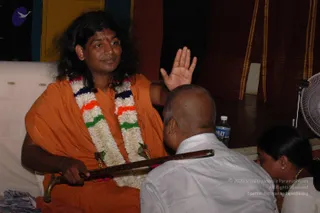 A long-haired man in an orange saffron robe and a multicoloured flower garland sits on a white cushioned seat, raising his right hand in blessing while holding a long wooden sword or staff with a brass hilt. Before him a bald man in a white shirt bows forward, and a woman with hair in a bun and gold earrings kneels at the right edge. A water bottle, a white floor fan and dark draped cloth stand behind them. The man's long dark hair, saffron robe and garland fit the Swamiji candidate cues, so the appearance-based label applies.