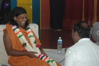 A photograph of a long-haired man in an orange saffron wrap smiling broadly while seated on a white cushioned seat. He wears a multicoloured garland of white flowers with orange, green, blue and pink accents and a red tilak on his forehead, and his hand rests on the garland. Facing him from the right is a bald man in a white striped shirt, seen from behind, with a plastic water bottle and a glass on a white table between them. A dark red curtain, yellow and blue panels and a white fan are in the background. The scene shows a sitting darshan exchange in a hall.