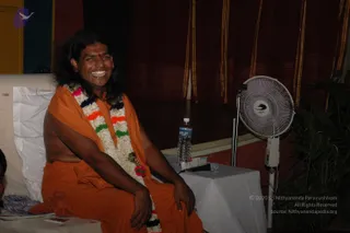 A smiling man with long dark hair sits on a white-cushioned seat wearing an orange saffron wrap draped over his body and a multicoloured flower garland with white, orange, green and blue blossoms. He is bare-chested beneath the wrap. A standing fan, a bottle of Bisleri-style water and a microphone on a white-draped table sit beside him, with maroon curtains and a potted plant behind. A small reddish toy lies at the lower left.
