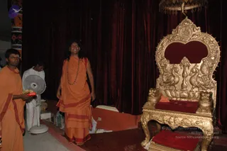 Indoor photo of a stage beside an ornate gilded throne with a red velvet seat and back panel, carved with peacocks and lion-like figures, under a dark red velvet curtain. Standing near the throne is a figure with long dark hair, a red tilak mark on the forehead, a rudraksha mala and an orange robe with red border. To the left, a man in an orange robe holds a tray of food. Behind him, a man in white clothing stands beside a white pedestal fan. A large plastic bag sits on the floor near the stage edge.