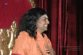 A photograph of a smiling man with long, dark, wavy hair seated on an ornate gold-carved throne with a red upholstered back. He wears a saffron-orange shawl over an orange garment, a white and red forehead mark, and a rudraksha mala draped across his chest, with one hand resting on his chest. Behind him hang deep red velvet curtains. a person, seated on the throne in saffron robes with rudraksha beads and long swept hair, as the page indicates his presence at the Deepavali event.