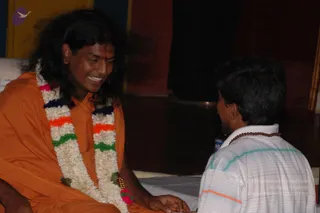 A seated man in an orange-saffron robe, with long dark swept-back hair and a multicolour flower garland of white jasmine with pink, orange, green and navy accents, smiles warmly while holding the hand of a younger man seated in front of him. The younger man wears a white and light-striped collared shirt and a brown rudraksha mala, seen from behind. The setting is an indoor hall with dark wooden flooring and a red and yellow wall. Swamiji (probable): the saffron robe, long swept-back hair and flower garland are the qualifying cues for the central figure, who appears to be receiving or giving blessings in the scene.