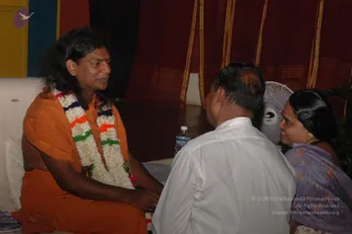 A seated man in an orange saffron robe with a white and orange-green tricolour-style flower garland faces a man in a white shirt whose back is toward the camera. Beside him a woman in a lavender floral sari with a gold earring sits smiling. A pale fan, a water bottle and red-and-gold curtains stand behind them. The seated man has long swept-back dark hair and a tilak, showing distinctive saffron robes and a garland, so he is a probable Swamiji candidate present in person.