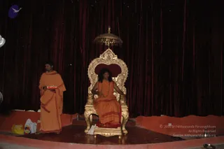 A photograph of an indoor stage with dark red velvet drapes behind it. A seated man with long dark hair wears an orange robe and a rudraksha mala, sitting on an ornate gold throne with a small gold canopy above it. To the left, a standing attendant in orange robes with red borders holds a red tray. Yellow and white plastic bags and a small metal lamp sit on the stage edge. The seated figure has distinctive long hair, orange robes and rudraksha beads, so he is an appearance-based a person candidate; the attendant is a standing priest-like attendant.