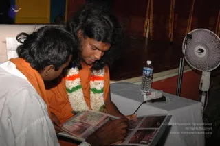 A photograph of two men seated at a table looking down at an open printed book or album with colourful pictures. The man on the left wears a white shirt with an orange shawl and bends toward the book, pointing at it. The man in the centre wears an orange-saffron robe with a white and orange-green tricolour-style garland of flowers, has long dark hair and a red mark on his forehead, and gazes at the pages. A water bottle, a microphone on a stand and a white-draped table sit in front of them, with a white electric fan at right and red and yellow striped drapes behind. The setting appears to be a hall with a stage area.