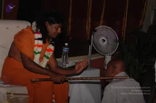 A photograph of a long-haired figure seated on a white-cushioned chair, dressed in saffron-orange robes with a multicoloured flower garland of white, pink, orange and green blooms around the neck. He holds a long wooden staff with brass-banded ends in one hand and extends the other palm toward a bald man in a striped collared shirt who is bent forward with his head lowered beside a small table. A water bottle, a white-draped table and a white pedestal fan stand nearby, with dark striped curtains and a potted plant behind.