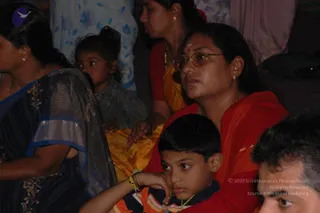 Photograph of a seated devotional gathering at night. In the foreground, a woman in a red sari with a red bindi and round rimmed glasses looks forward attentively, a young boy in a striped orange and navy shirt rests his chin on his hand beside her. To the left, a woman in a dark blue and gold patterned sari sits near a toddler in grey, and another woman in a yellow-orange sari is seen behind them. A man's partial face appears at the lower right edge. Background shows hanging fabrics. Several people are visible.
