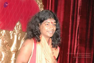 A person with long, dark, wavy hair stands beside an ornate gold throne with a red cushioned back, in front of deep red velvet drapes. The person wears a light beige shawl over a red garment, a red bindi and a tilak on the forehead, and a gold-coloured beaded necklace.