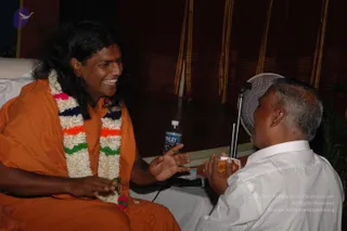 A photograph of two men seated indoors in conversation. On the left, a man with long swept-back dark hair wears an orange-saffron robe and a multicoloured garland of white flowers with blue, orange, green and pink bands, plus a rudraksha-style mala; he is smiling broadly and gesturing with his hand. Swamiji (probable) is indicated by the long hair, saffron robe, garland and mala cues. On the right, a man with short greying hair in a white shirt sits with his palms joined, holding a small sweet or offering. Behind them are dark red drapes with gold stripes, a white table with a water bottle, and a standing fan.