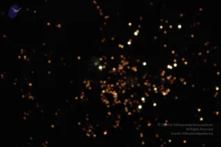 A night photograph of a firework display against a black sky. Many small orange and white glowing sparks radiate outward from a central burst, with scattered bright points drifting across the frame. No people, figures or ritual objects are visible.