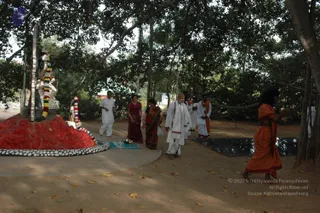 A photograph taken outdoors under a large banyan tree at an ashram grounds. On the left, a red-painted mound with a black-and-white bordered base holds a dark deity murti decorated with a multicoloured garland and a metal figure. A man in white shirt and dhoti walks in the middle distance. In the centre, a bald-headed man in white robes with a red-bordered shawl walks on a sandy path, accompanied by a woman in a maroon sari and another woman in an orange-and-brown sari. On the right foreground, a figure in an orange saffron robe with long dark hair walks away, carrying an object. Other people in white and orange clothing stand behind, and a dark mat lies on the ground near the tree trunks.