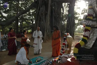 Outdoor scene beneath a large banyan tree with many aerial roots. A man in a white shirt and white dhoti kneels on a green cloth at lower left, tending a small brass lamp and oil dish. Standing at centre, a bald man in white kurta and dhoti with a white shawl holds his hands together in prayer. To his right, a woman in an orange sari wearing a mala stands with hands folded. At left, a woman in a maroon sari holds a small white bag, and another woman in an orange sari stands nearby. At right, a deity murti with a golden hooded cobra is adorned with garlands of red, yellow and green flowers, with a bell-metal kalasha-like silver base. Several more people sit in the background.