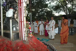 Outdoor ritual at a large black stone deity murti under a banyan tree, its base smeared with red and wrapped with a striped garland. On the right, a woman in a bright orange-and-red sari stands with hands folded in namaste, and a man in a white dhoti with a white shawl and orange-red border, a shaved head and white beard holds a lit oil lamp for aarti. Beside them, a man in an orange shawl and white dhoti, another in white, and a woman in a pink-magenta sari stand together. Several people in white are visible in the background. A small brass bowl and a blue mat with puja items sit on the ground near a circular stone border.