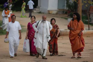 A photograph of five people walking along a dirt path outside a white building with trees and shrubs nearby. On the far right, a man in an orange saffron robe with long dark hair and a rudraksha mala walks barefoot-style in sandals, holding beads in one hand; the mala and swept-back hair are the cues. Beside him is a man in a white kurta and dhoti with a white shawl over his shoulder, bald with a white beard, carrying nothing. Next to them a woman in a magenta sari carries a white shopping bag, a woman in a rust-orange sari carries a bag over her shoulder, and an older man in white kurta and white dhoti walks at the left holding a small bag. Other people stand in the background, including a figure in an orange shawl and white clothes.