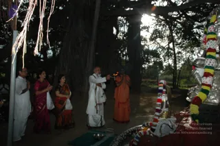 Outdoor evening scene beneath a large banyan tree at dusk. A bald, bearded man in white dhoti and shawl holds a flaming lamp or aarti flame toward a silver-coloured deity murti decorated with multicoloured garlands. Beside him a saffron-robed sannyasi with his hands joined stands in devotion. To the left, three devotees stand with hands folded: a man in white, a woman in a magenta sari, and a woman in an orange-and-black sari, one holding a white bag. Items on a green mat and a silver plate sit in front of the murti, a red-and-white garland draped over the base. Hanging white threads and tassels frame the upper left.