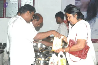 A photograph of an indoor food-serving scene at an ashram event. In the foreground, a man in a white shirt and glasses stands at a counter lined with stainless steel bowls filled with food, reaching toward a bowl. Beside him, an older man in white clothing pours or handles food. A woman in a white outfit with a red handbag and gold bangles holds open a yellow-and-white packet, receiving food. Behind them, a younger person in a white t-shirt and another person in red stand near a wall. A large portrait poster on the right shows a smiling man with dark hair in white clothing, partly cropped. Plastic carry bags sit on the counter.