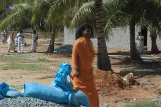 A woman with long dark wavy hair in a saffron-orange robe stands on a dirt construction site beside a pile of reddish earth and blue woven sacks of gravel. Tall coconut palms and a whitewashed brick building frame the scene. Several people in white and cream clothing walk or stand in the background, including a man in white trousers and shirt and women in white saris with borders. The woman appears to be a person by appearance cues of long swept hair and saffron robe, though this is uncertain given the distance and framing.