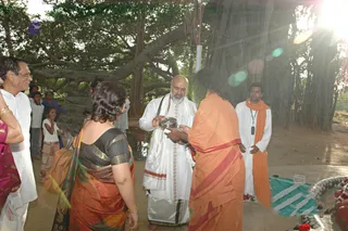 Outdoor photo beneath a large banyan tree with hanging aerial roots and sunlight flaring through the canopy. A bald, bearded man in white dhoti and shawl, with a white sacred thread, holds a silver plate toward a person in saffron-orange robes with a red-edged cloth, whose back is to the camera. Nearby, a woman in an orange and black sari with gold-trimmed borders stands with her back to the camera, and a man in white with glasses and a white shirt stands at left. A young man in white with an orange scarf and identification lanyard stands at right. Children and a few other people are visible in the background near the tree. In the lower right, a green-and-white mat, a glass bowl and red flowers sit on a raised platform. Appearance cues for the saffron-robed figure are limited, so the identity is uncertain.