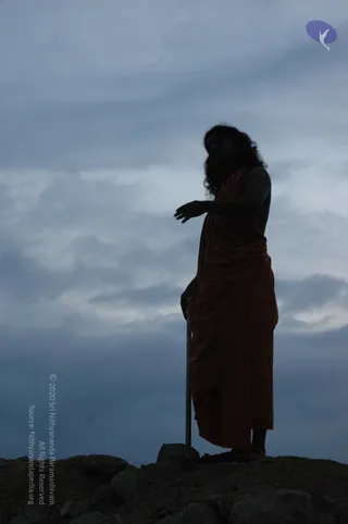 A silhouetted figure in a reddish-orange wrap stands on a rocky outcrop under a dim, cloudy blue-grey evening sky. The figure has long, loose, wavy hair and a full beard, with one arm extended outward and the other hand resting on a tall metal staff or rod planted in the rock. Tones are muted, with the orange cloth the only warm colour against the cool sky. A small purple logo appears in the top right corner and a vertical copyright credit runs along the left edge. The figure has long wavy hair, a full beard, an orange wrap and a tall staff, which matches the appearance-based cues for a person as a person in ascetic dress, though the silhouette makes this uncertain.