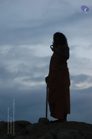 A photograph at dusk of a lone figure standing on a rocky outcrop against a grey-blue cloudy sky. The figure wears a long reddish-orange robe draped over one shoulder and holds a tall slender staff in one hand while raising the other hand near the chest. Long dark hair falls past the shoulders. The figure is in silhouette, with rocks in the foreground and a small purple logo in the upper right corner.