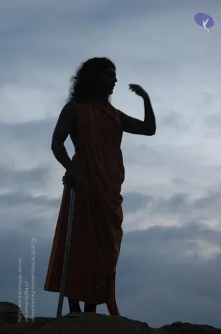 A photograph at dusk of a silhouetted figure standing on a rocky ridge against a grey, cloudy sky. The figure wears a rust-orange or saffron wrap draped over the body, with long, wavy hair and one arm raised with the hand near the face. The other hand rests at the hip, and a long metal staff or rod stands beside the figure. A small purple logo appears in the upper right corner.