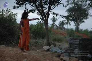 Outdoor photograph of a long-haired man in an orange saffron wrap, bare-chested and shod, standing on a dirt ground with a slender walking staff in his left hand while his right arm extends to point toward the left. He is beside a leafy tree, with a low stone wall of rough-cut blocks and scattered boulders to the right, overgrown shrubs, palm trees and a grey overcast sky behind. No other people are visible.