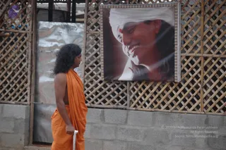A photograph of a long-haired man in a bare-shouldered orange robe walking past a bamboo lattice fence beside a large printed portrait of a smiling man with a white head wrap and a red mark on his forehead. The walker holds a white cloth in one hand and walks along a concrete block wall, with a silver sheet beside him. The portrait is mounted on the lattice and is not a physical person.