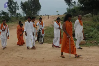 A group of devotees walks along a dirt road lined with green shrubs and trees under an overcast sky, a motorcycle parked among them. In the foreground a figure with long dark hair wears a plain saffron-orange robe and sandals and holds a tall silver walking staff. Around him, men in white kurtas and dhotis, some with saffron-orange shawls draped over their shoulders, walk barefoot or in sandals. Two women in orange saris walk on the left. Further up the road, two small distant figures are visible on the hill.