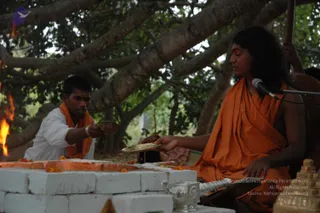 Outdoor homa ritual beneath a large spreading tree. A fire burns in a brick-lined fire pit on the left, with marigold petals scattered on the bricks. A young man in white shirt and orange shawl sits at the fire pit holding a small offering. Seated on the right, a figure in orange saffron robes with long swept-back hair and a tilak holds a brass plate of offerings toward the fire; a microphone stands before him. A silver kalasha-style vessel and a gold deity murti sit in the foreground. Swamiji (probable) is shown seated in saffron robes with long hair and tilak, officiating the homa.