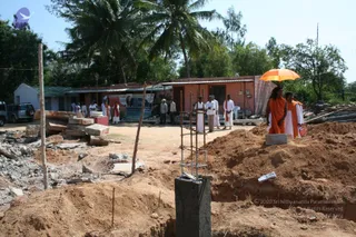 A photograph of a construction site with excavated red earth, rebar cages standing in concrete footings, scattered stone slabs and rubble in the foreground. Behind it stand a row of low buildings with corrugated roofs and a palm-lined backdrop. A group of about six people in white clothing and saffron-orange robes stands near the excavation; one woman in orange holds a bright orange umbrella. A large printed banner is attached to the building behind them.