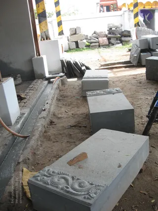 A stone yard or workshop outdoors with several grey granite blocks lying on bare dirt. The front block has carved scrollwork along its edge, and two more carved blocks sit behind it. A thin trench of wet concrete runs along the left, with a hose beside it. Stacks of rough stone and dressed slabs are piled against a white wall in the background, and black-and-yellow striped safety posts stand at the left. No people are visible.