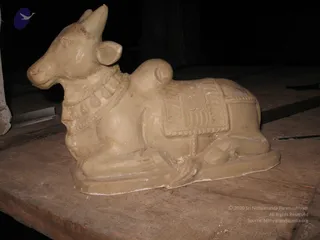 A pale, unpainted plaster or clay sculpture of a recumbent bull, the Nandi, lies on a rough wooden board in a dim room. The bull has an upturned head, a decorated collar with round medallions, and a patterned saddle cloth across its back. No people are visible. The base is a smooth raised plinth, and the surface shows small chips and marks typical of an unfinished or freshly cast figure.