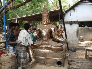 A large polished bronze deity murti seated in lotus-style posture on a metal-fronted base with drawer panels, under a corrugated roof beside a whitewashed wall outdoors among palm trees. The murti has a tall ornate crown, multiple arms and heavy jewellery, and a hand holds a trident-like implement. Several workers in lungis and shirts, one bare-chested, stand around it, one adjusting the figure with his hands. A sack of material sits in the foreground. The image shows a craftsman workshop setting rather than a ritual. No identifiable a person candidate is clearly visible.