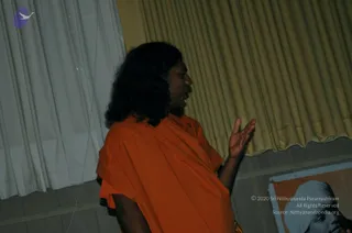A person stands in profile on a low stage in front of pale yellow and white curtains, speaking with one hand raised open in a gesture. The person wears a loose orange-saffron wrap over the shoulder and has long dark, swept-back hair. At the lower right, a framed printed portrait shows a figure in a light head covering against an orange background. The room is dimly lit.